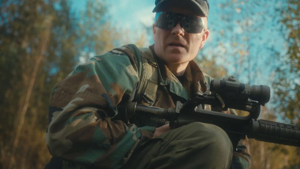 Still from a video, where a character is kneeling and looking down on the camera. He is holding the Green Diamond Carbine in his hands.